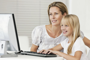 Young girl operates a computer while her mother watches on. Horizontal shot.